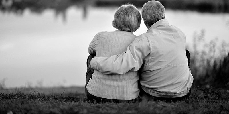 Backs of retired couple sat on grass overlooking a pond